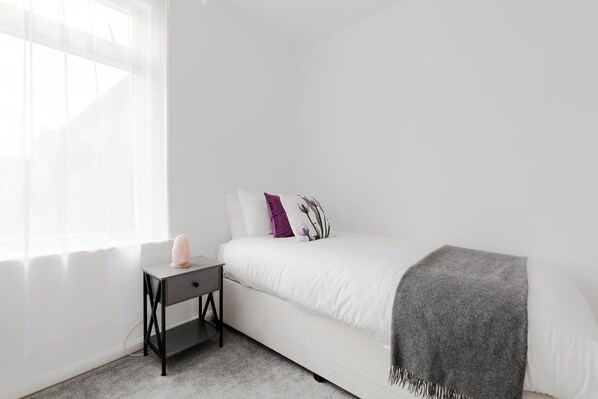 A minimalist bedroom with a white bed, a small bedside table, and a window with sheer curtains. - 4 Bed House - Sleeps 6 - Garden, Pets, Wifi (London)