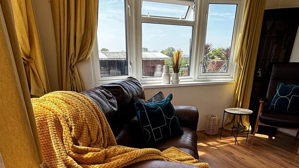 A cozy living room with a brown leather sofa, a woven side table, and a view of the outside through large windows. - 2 Bed Chalet on Working Farm (Cullompton)