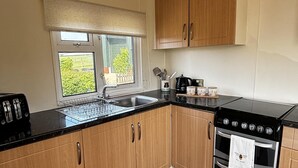 A kitchen with wooden cabinets, a black countertop, a window with a view, a stove, and a sink. - 2 Bed Chalet on Working Farm (Cullompton)