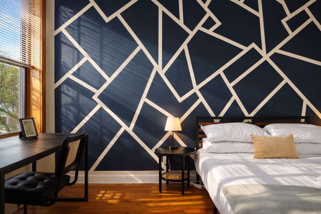 Ein Zimmer mit Wand in geometrischem Muster, einem Bett mit weißen Leinen, einem Nachttisch mit Lampe, einem Sessel und einem Schreibtisch mit Bilderrahmen.