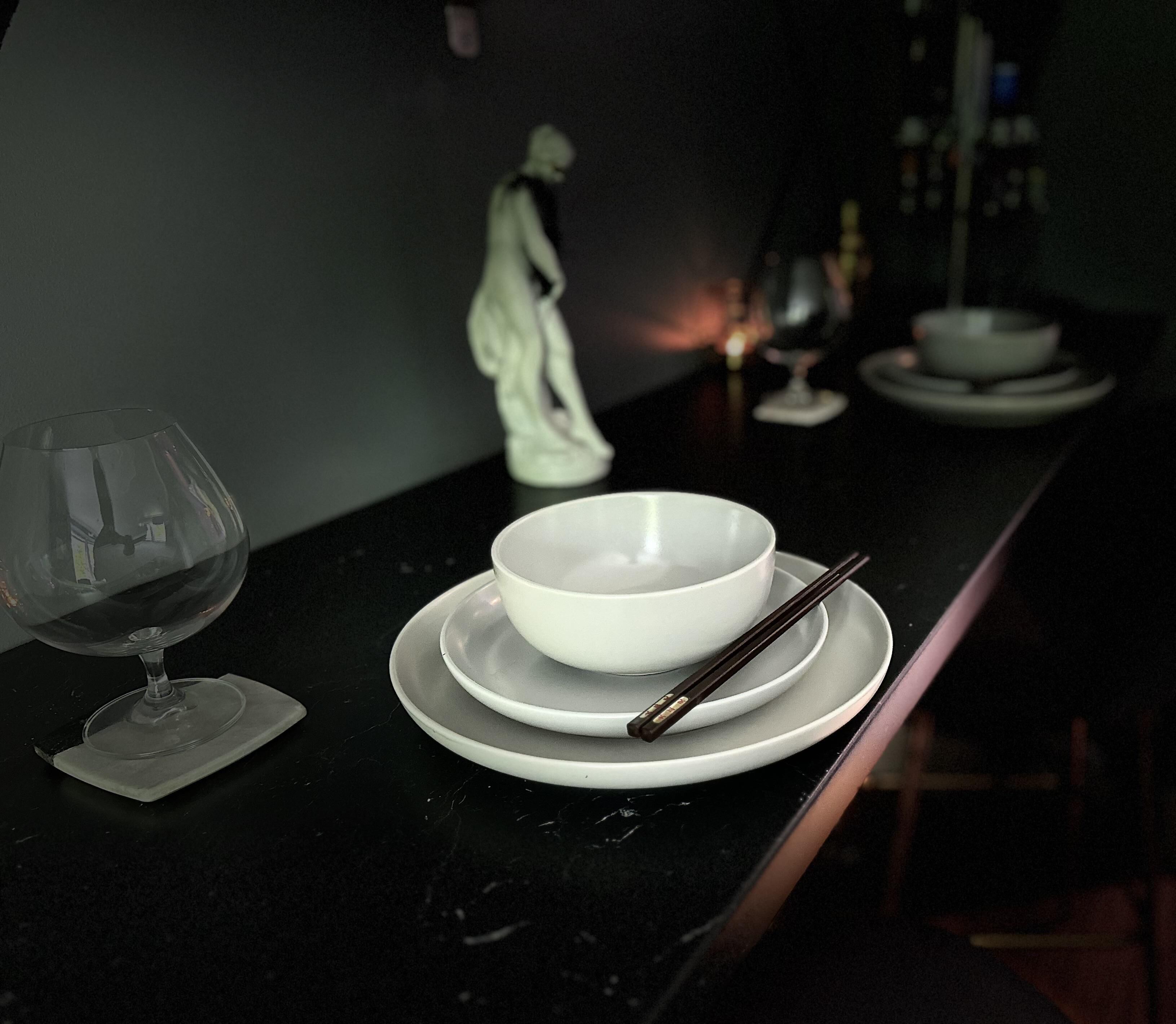 A dimly lit room with a dining table set for two. A wine glass and chopsticks are on the table, and a statue is in the background.