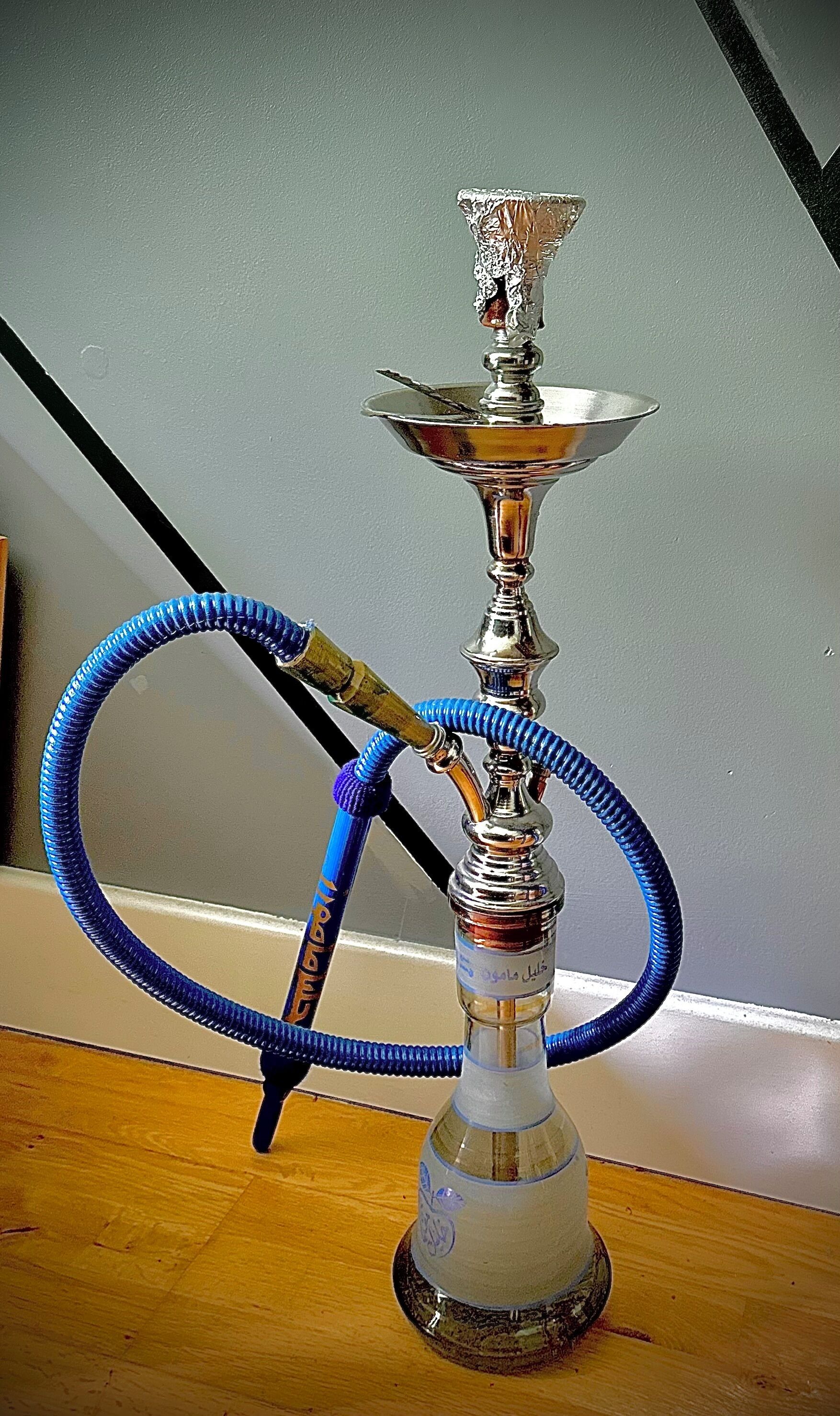 A hookah with a blue hose and a silver base, placed on a wooden surface against a grey wall.