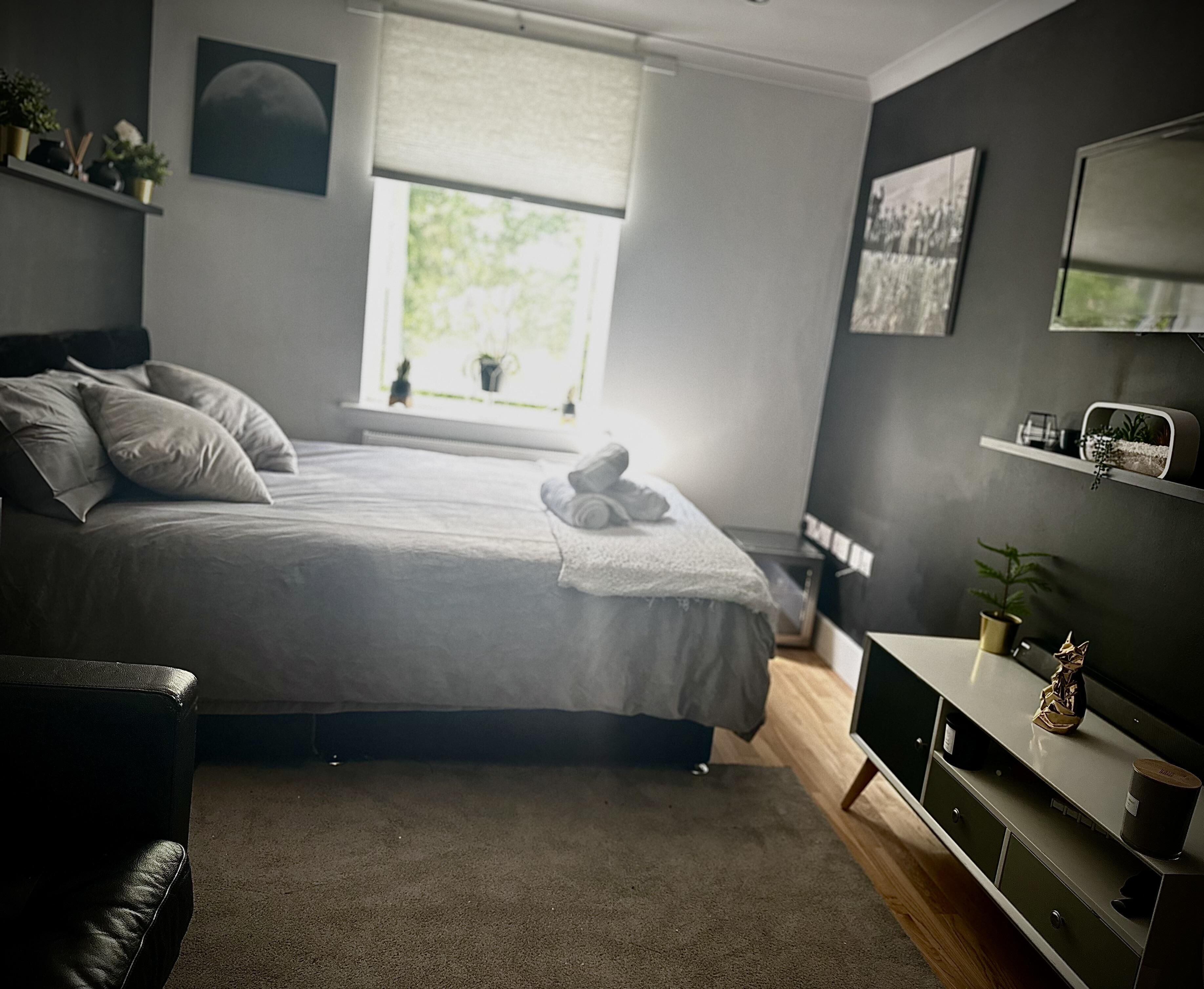 Ein Schlafzimmer mit einem Bett, einem Nachttisch, einer Kommode, einem Fenster mit Jalousien und einem Regal mit Pflanzen.