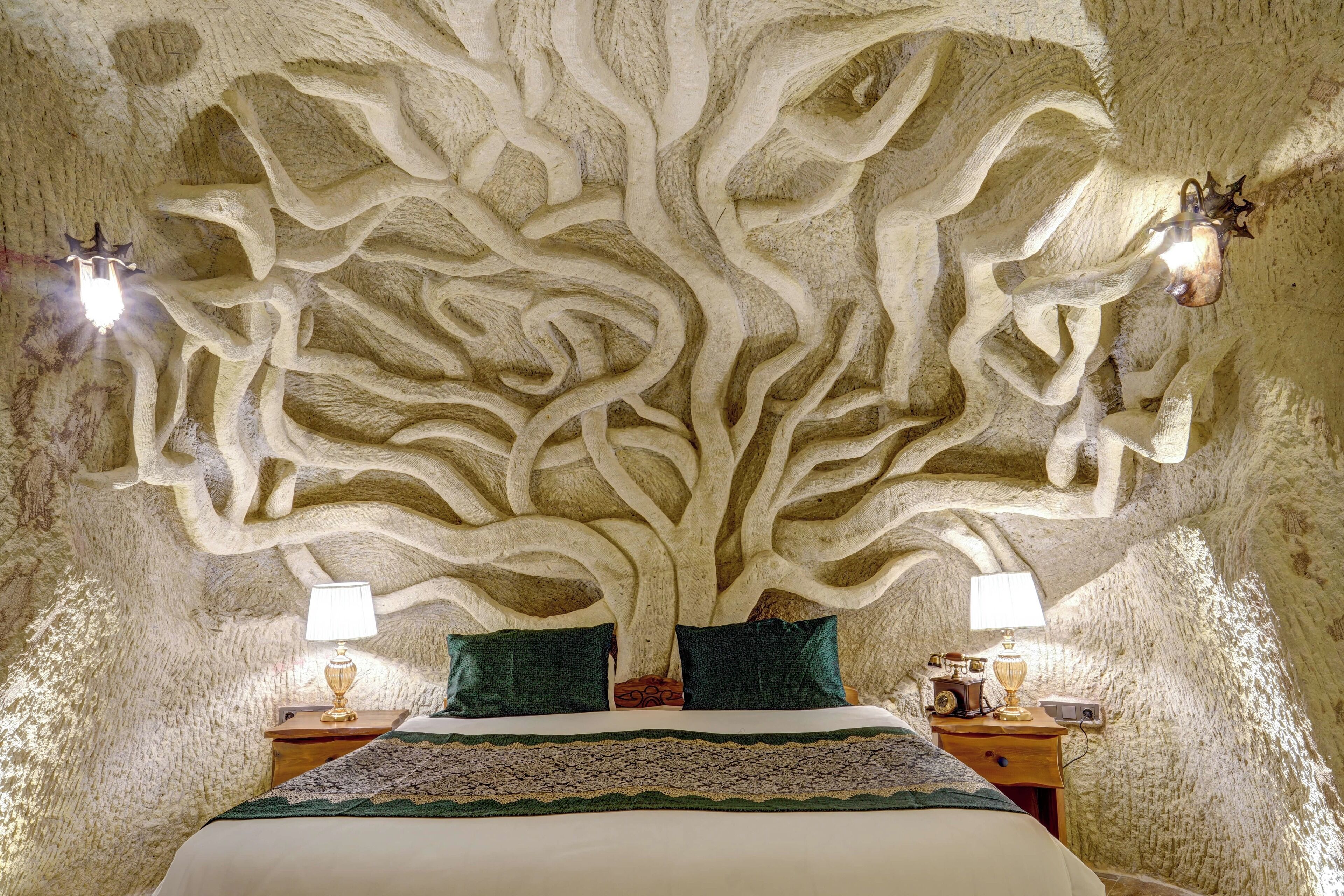 Un dormitorio con una pared tallada que evoca un árbol, dos mesitas de noche con lámparas y una cama con colcha verde y blanca.