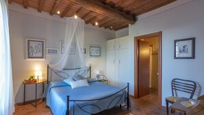 Un dormitorio con cama, mesitas de noche, una silla, un armario y una puerta que da a otra habitación.