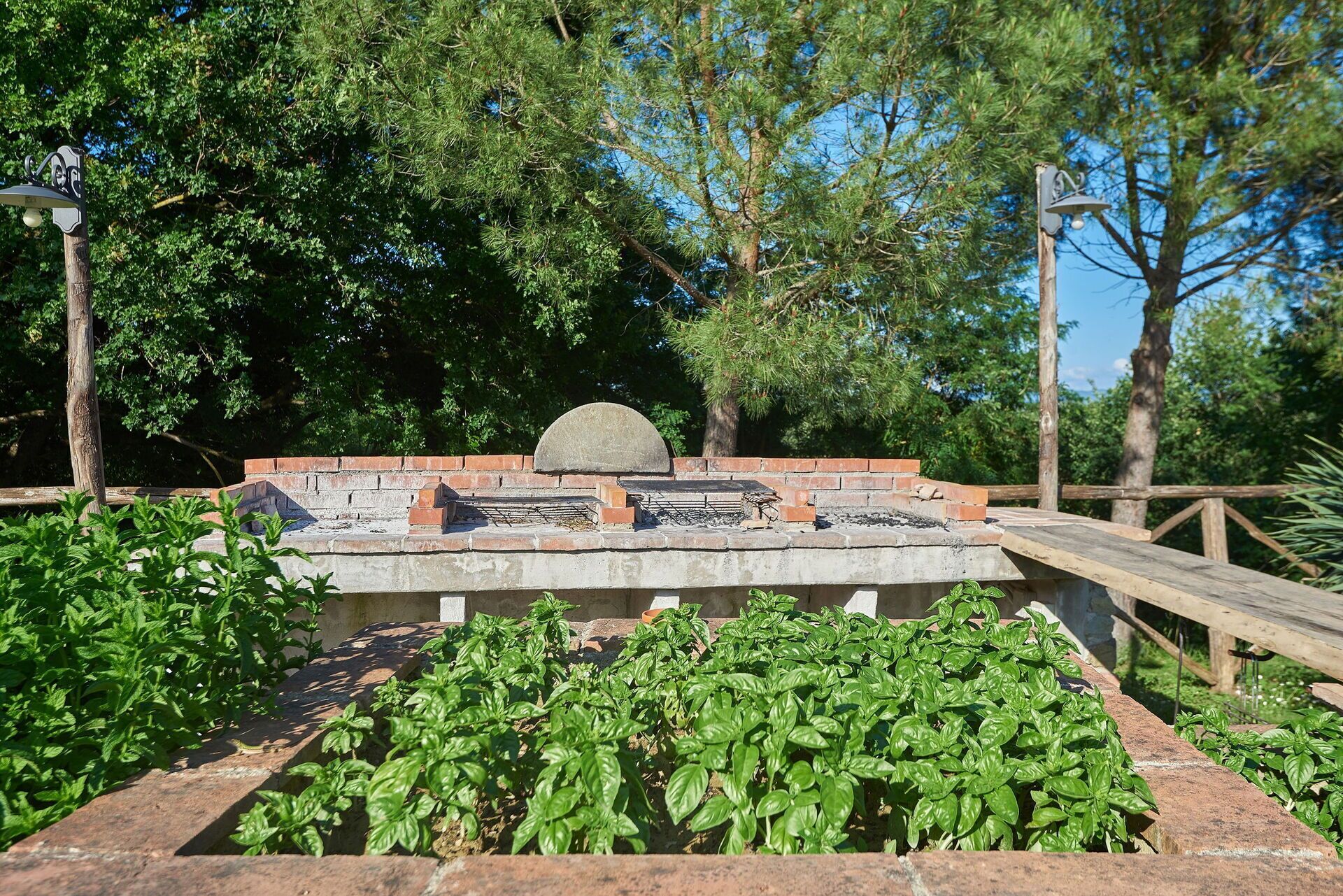 A brick barbecue grill with a stone top, surrounded by greenery and trees.