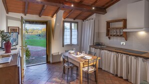 A cozy kitchen with a wooden table and chairs, a window with curtains, and a view of a green landscape. - Le Fornaci - Apartment with pool (Latereto)