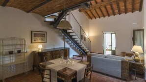 A dining area with a table set for four, a staircase, and a sofa in a rustic room with exposed brick walls and wooden beams. - Le Fornaci - Apartment with pool (Latereto)