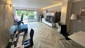 Ett modernt vardagsrum med ett fiskbensmönstrat golv, en glasvägg, en soffa, en tv och en matplats.