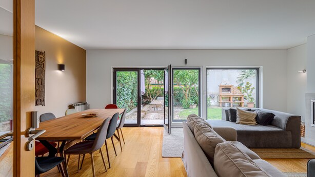 A modern living space with a wooden dining table, a sofa, and a sliding glass door leading to an outdoor patio. - Casa das Fontes – House of Wells with Mountain View, Private Garden and Wi-Fi (Parede)