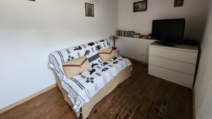 Une petite pièce agréable et chaleureuse, avec un plancher en bois, un canapé blanc orné de coussins à motifs, un bureau blanc avec un écran d’ordinateur et un luminaire au plafond.