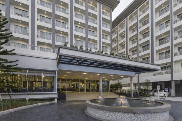 Un hotel de varios pisos con entrada cubierta, una fuente central y un área de estacionamiento.