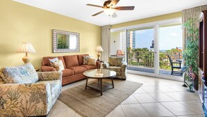 A living room with a sofa, armchair, and coffee table, a ceiling fan, a large window with a view of buildings, and a balcony with chairs. - Walk 2 Beach~Spacious Balcony~100ft2Bch~Fab Lagoon Pool~Sterling Shores 307 (Destin)
