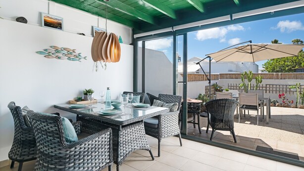 Un comedor con sillas de mimbre y una mesa con cubierta de vidrio, un techo con vigas verdes y vistas a un patio exterior con mesa y sombrilla.
