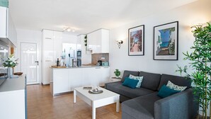 Una sala de estar moderna con un sofá gris, una mesa de centro blanca y una cocina al fondo.