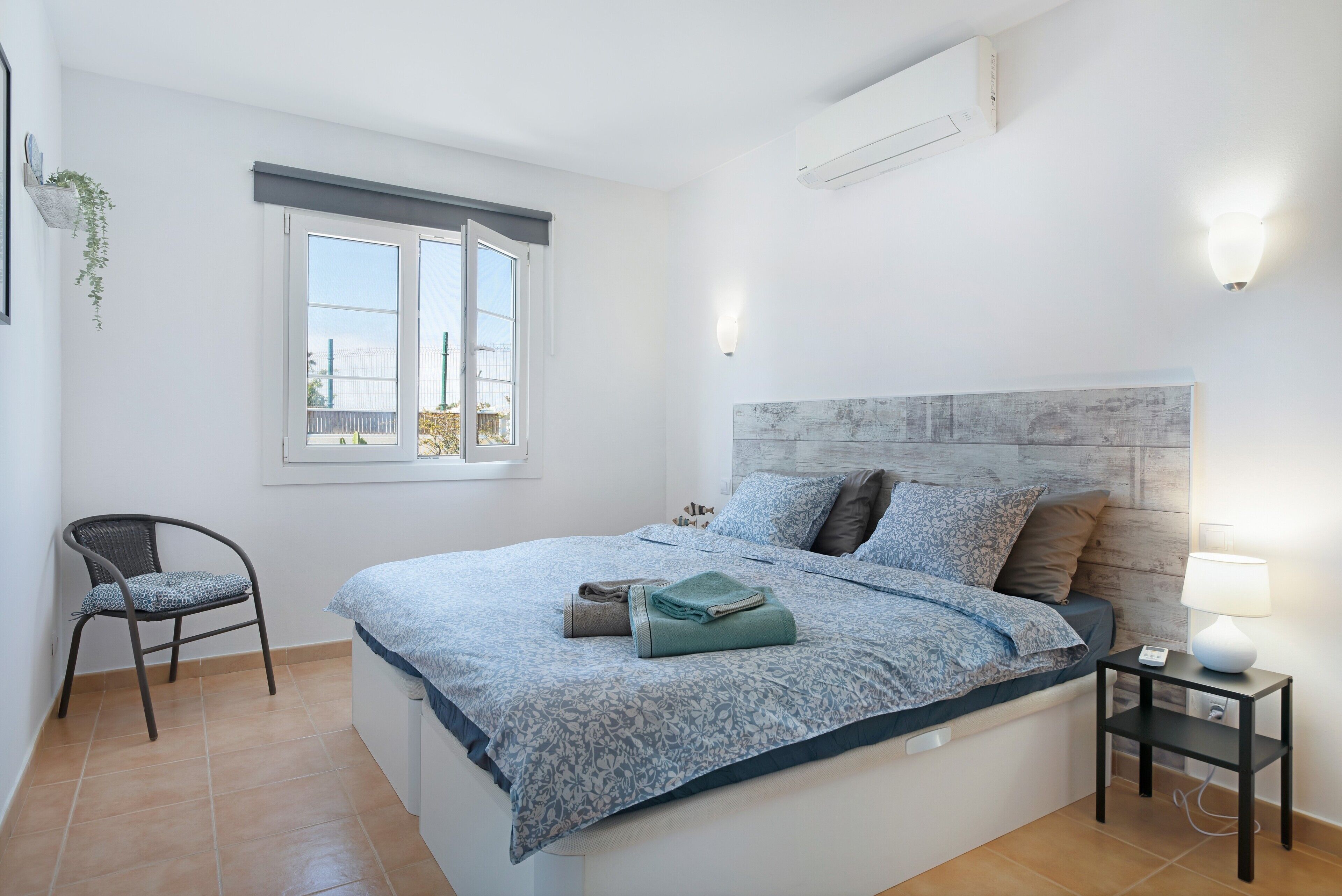 Un dormitorio con cama, una silla, una mesita de noche con lámpara, una ventana y un equipo de aire acondicionado.