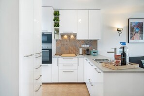Une cuisine moderne avec des armoires blanches, un four encastré, une machine à café et une planche à découper en bois.