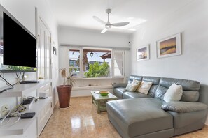 A living room with a grey sofa, a television, a ceiling fan, a window with a view, and a potted plant.