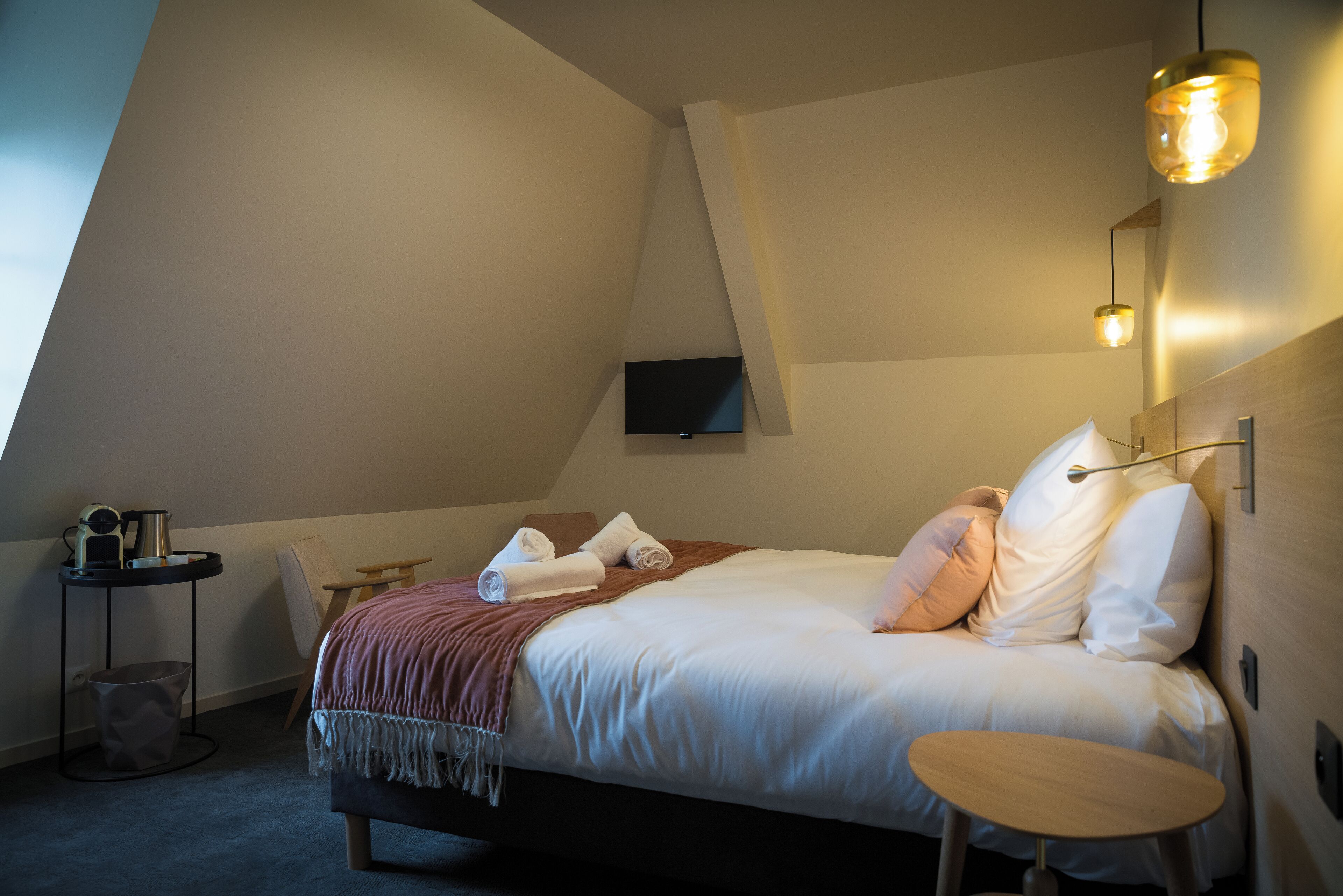 A modern hotel room with a bed, a small table, a TV mounted on the wall, and a unique sloped ceiling.