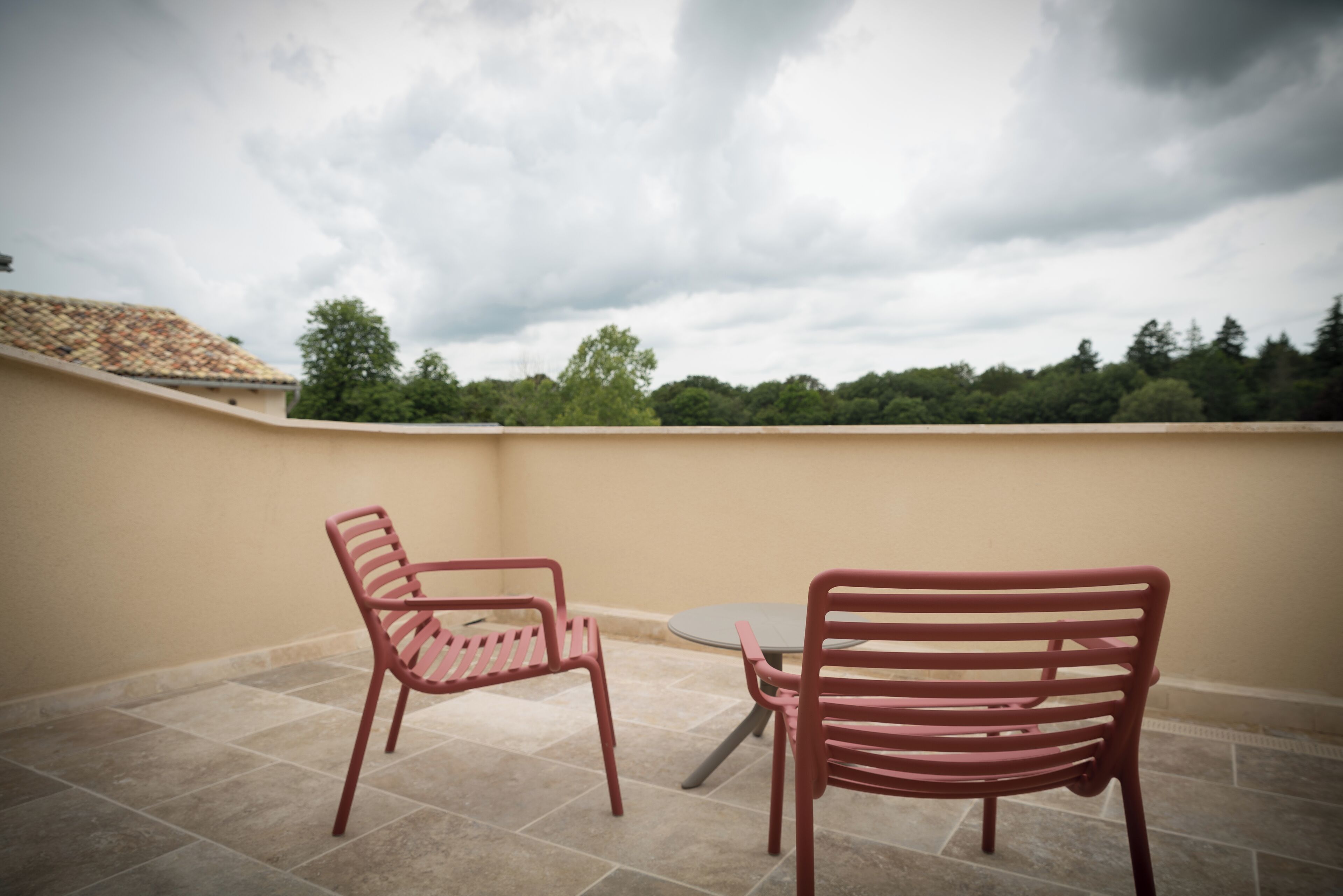 Una terraza con dos sillas de metal y una pequeña mesa, rodeada de paredes color beige y piso de baldosas, con vistas a un área boscosa bajo un cielo nublado.
