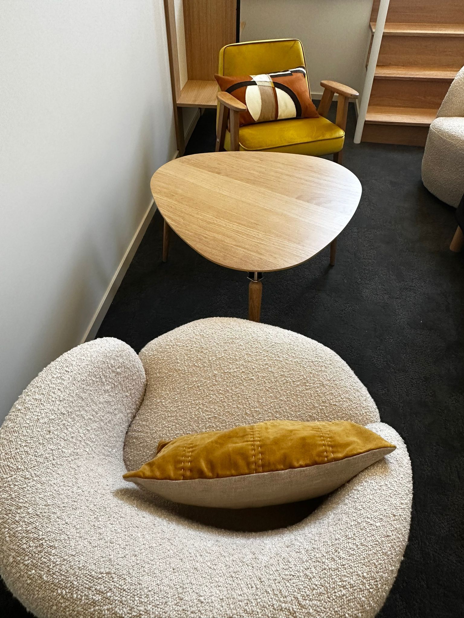 Une pièce moderne avec un canapé rond de couleur beige, agrémenté d’un coussin moutarde, une table en bois et une chaise jaune avec un coussin à motifs.