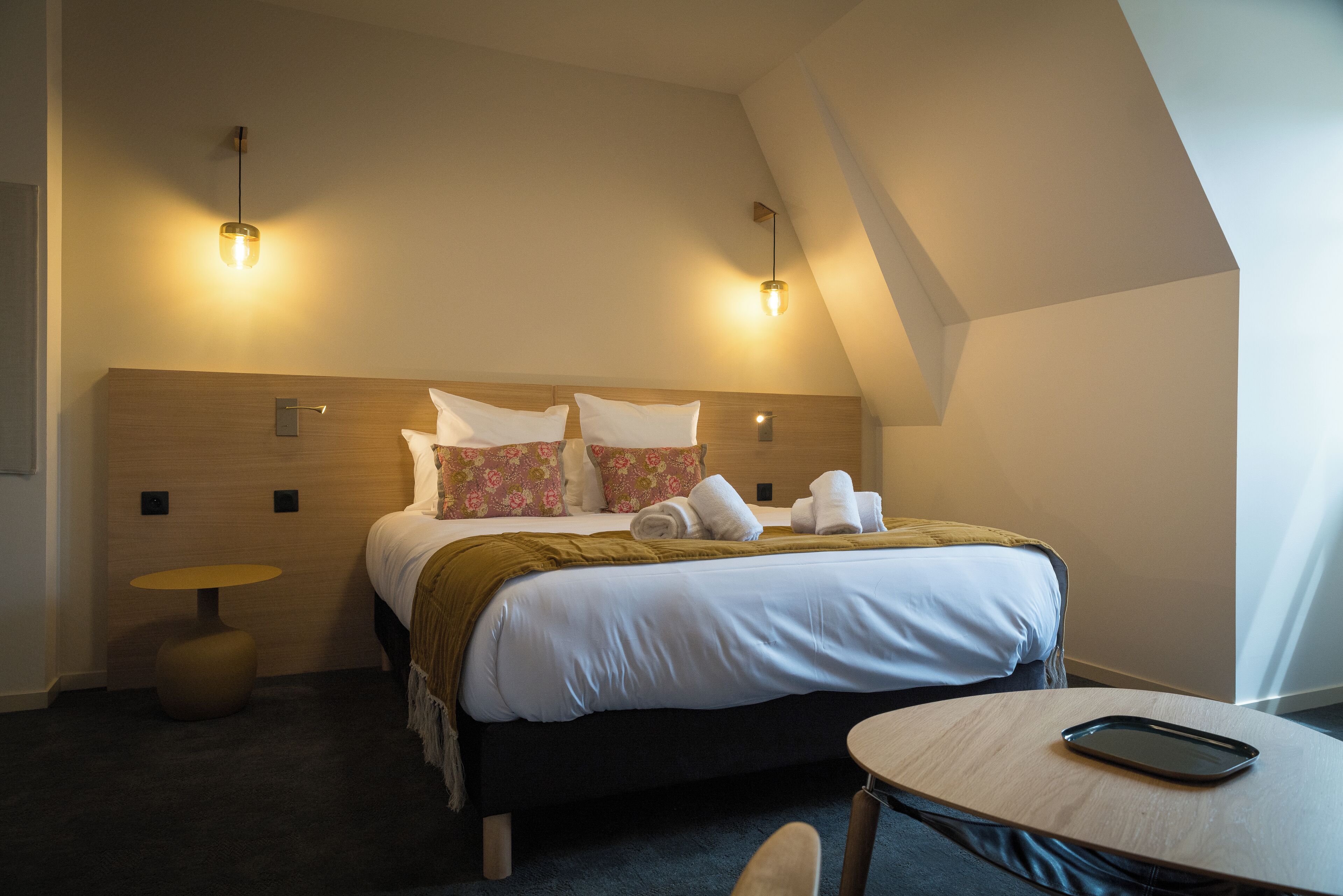 A hotel room with a bed, two bedside tables, a round table, and a lamp.
