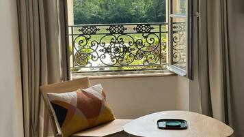 A room with a wooden chair and table, a view of a balcony with a metal railing, and a window with curtains.