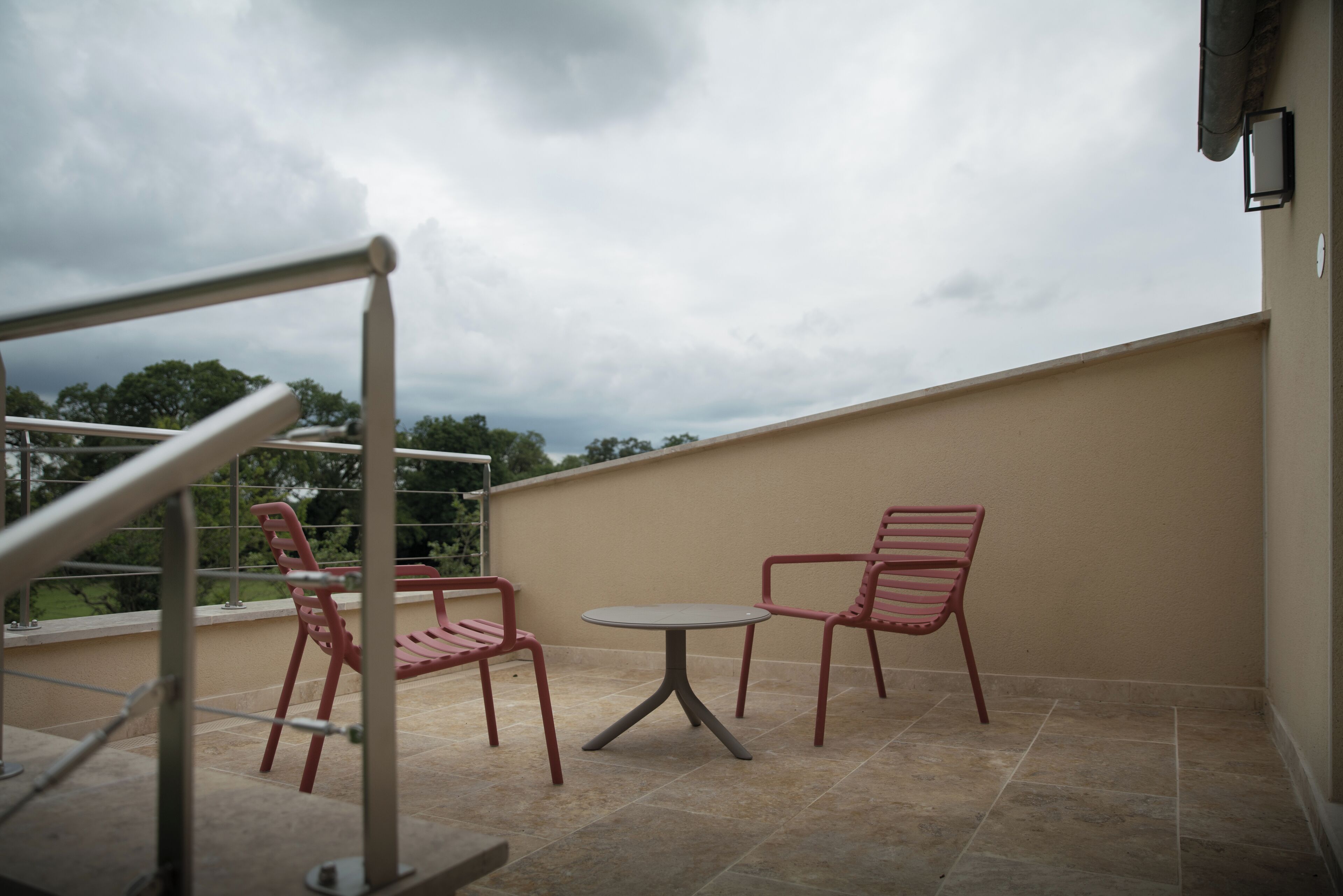 Un balcon avec deux chaises rouges et une petite table, donnant sur un jardin et un ciel nuageux.