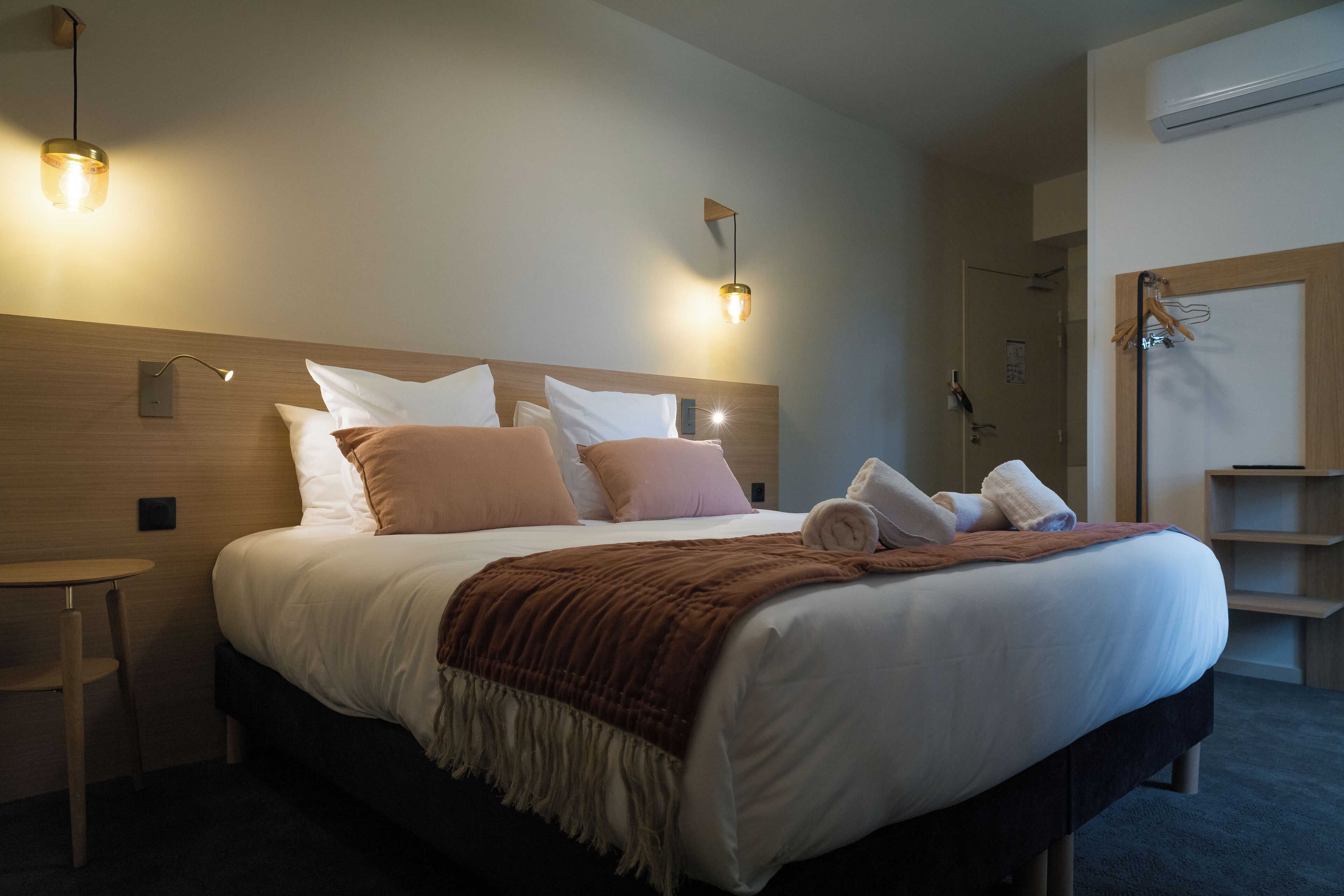Una cama bien hecha con una manta marrón, dos lámparas de noche y una mesita de noche de madera.