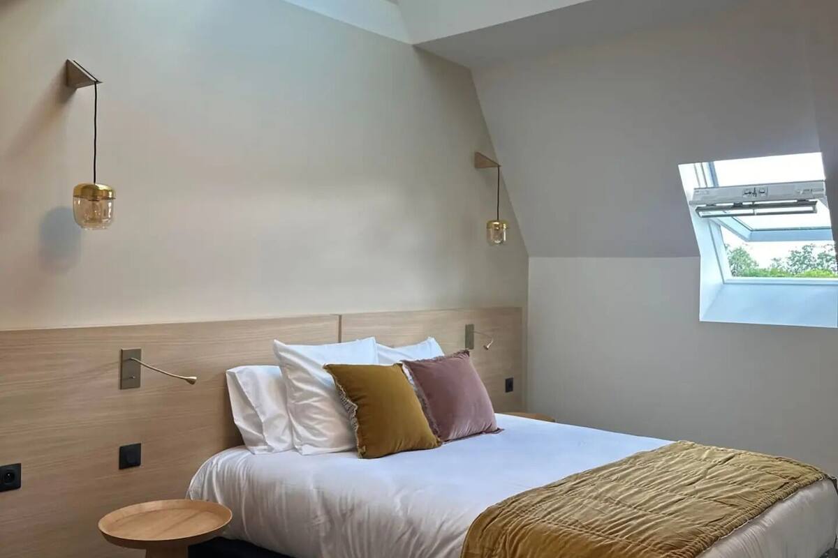 Un dormitorio moderno con tragaluces, una cama con muchos cojines, una mesita de noche y una ventana.