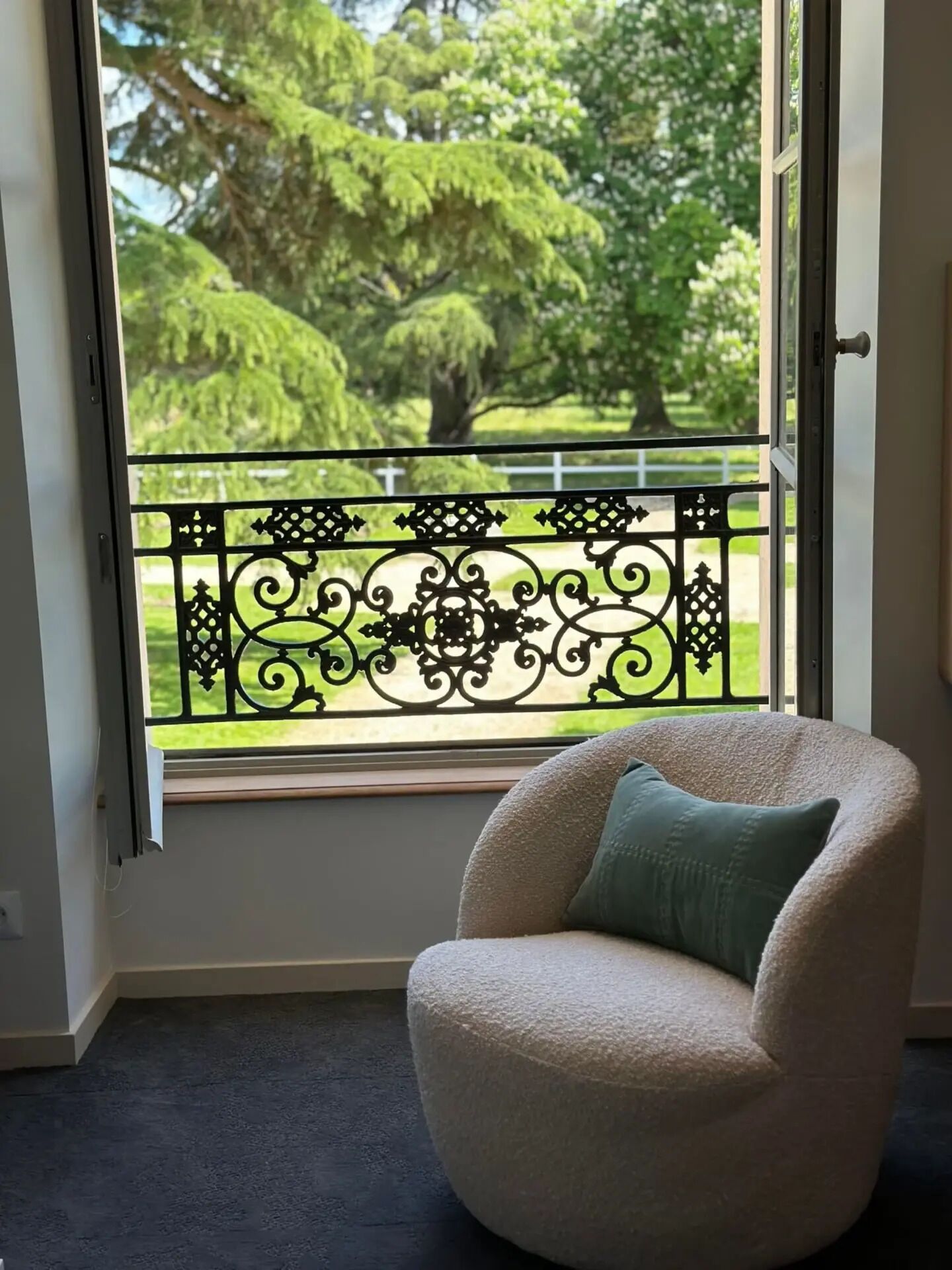 A room with a view of a garden through a window with a decorative black metal railing.