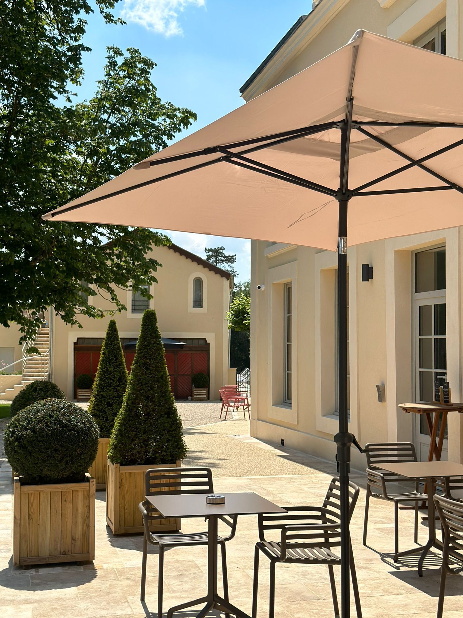 Um pátio externo com mesa e cadeiras sob um grande guarda-sol, cercado por arbustos bem aparados e um edifício com fachada bege.