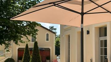 Um pátio externo com mesa e cadeiras sob um grande guarda-sol, cercado por arbustos bem aparados e um edifício com fachada bege.