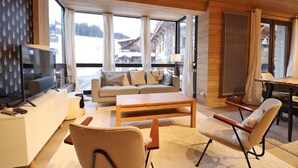 Una sala de estar moderna con sofá, mesa de centro y sillas, televisor y ventanas grandes con vistas a edificios y árboles.