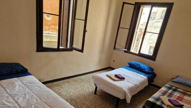 A room with two beds, one with a blue pillow and the other with a white pillow. There is a small table with a plaid tablecloth and a purple towel on it. - 2 bedroom apartment in venice (Venezia)