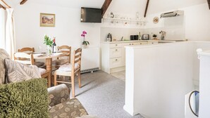 Eine gemütliche, gut beleuchtete Küche mit Holzbalken, ein Essbereich mit Tisch für Zwei und ein Wohnzimmer mit Sofa und grüner Decke.