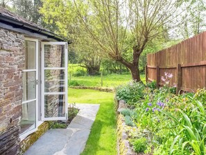 A stone building with a glass door, a stone pathway, a wooden fence, and a garden with various plants and flowers. - Hingham Mill Cottage (Wadebridge)