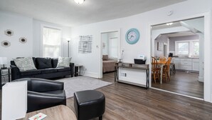 A modern living room with a black leather sofa, a wooden coffee table, and a black leather ottoman. There is a dining area with a wooden table and chairs, and a kitchen visible through an open doorway. - 3 bed/1 bath lower unit conveniently located in Lakeside. (Muskegon)