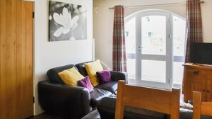 A living room with a black leather sofa, wooden dining table, and a television on a wooden cabinet. - 2 bedroom accommodation in Netherton Near Rothbury (Netherton Near Rothbury)
