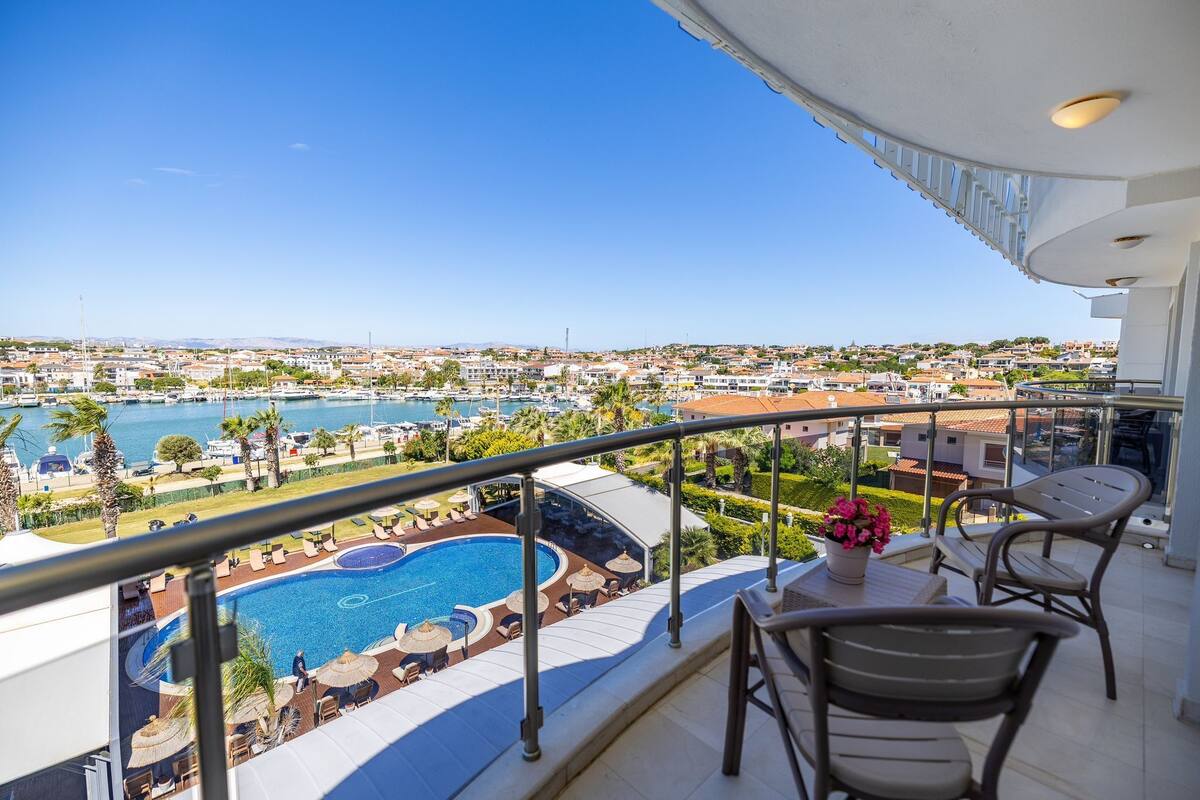 A balcony with a pool, chairs, and a table overlooking a marina and cityscape.