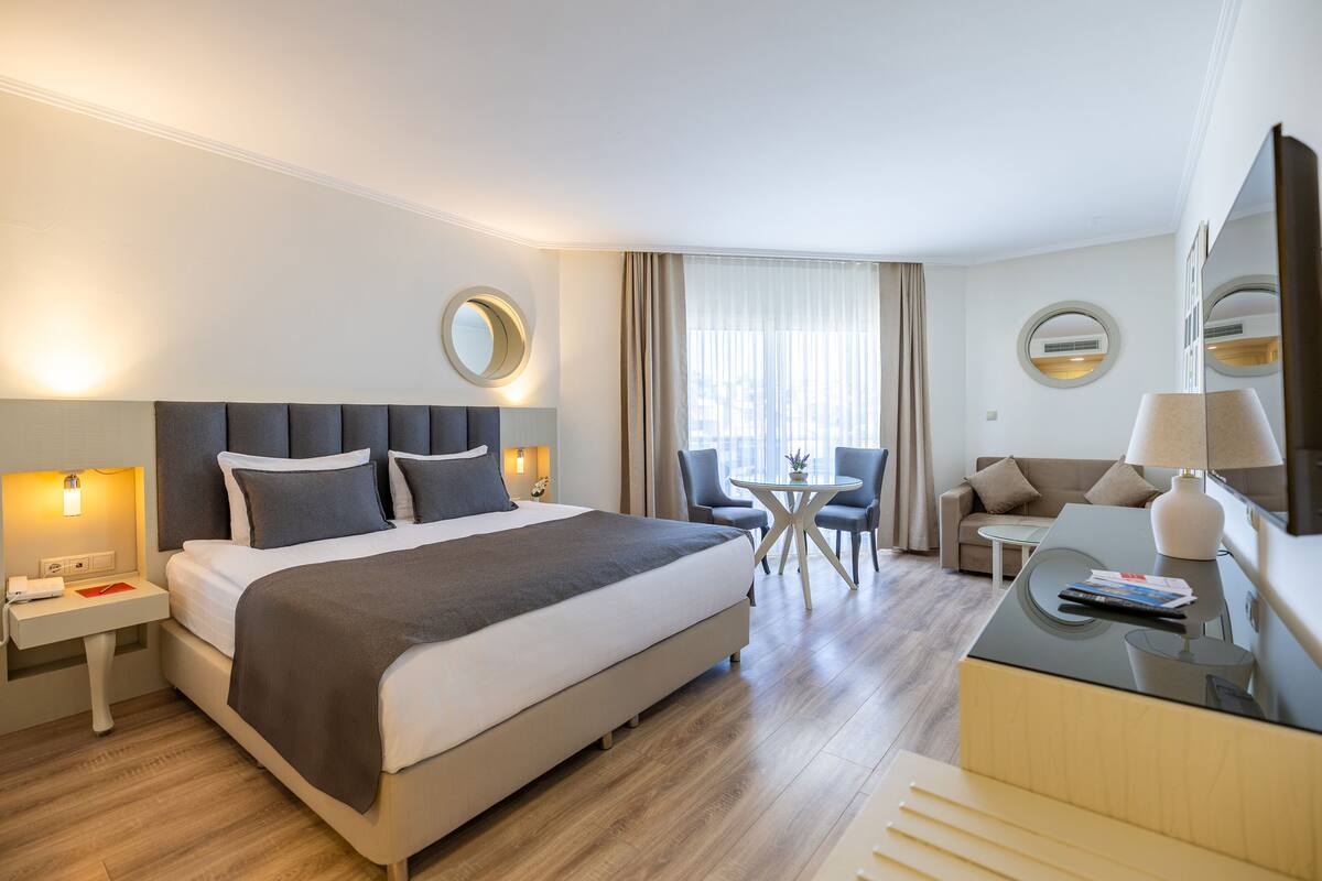 A modern hotel room with a large bed, bedside tables, a round mirror, a small round table with chairs, a sofa, and a TV.