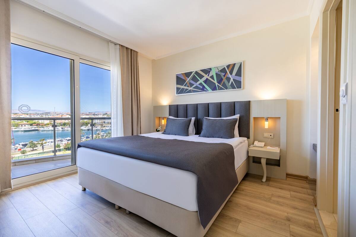 A modern hotel room with a large bed, a nightstand, a wall-mounted lamp, and a view of a marina.