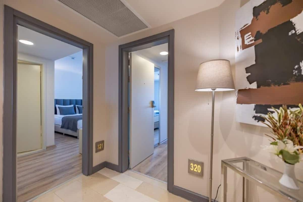 A hotel corridor with a bed in the adjacent room, a lamp, and a wall-mounted number plate.