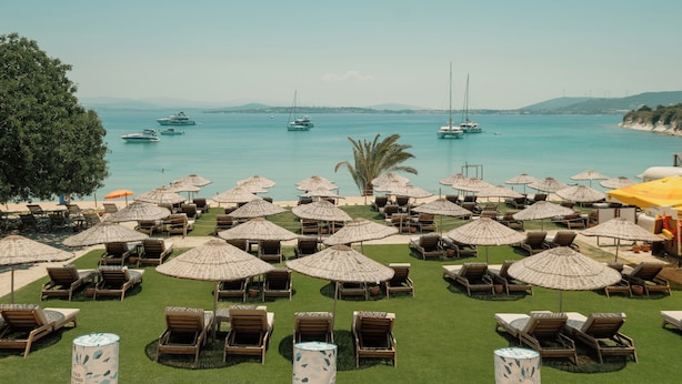 A beachside lounge area with multiple sun loungers and umbrellas, a clear view of the sea with boats, and a distant view of hills. - Sisus Marina Hotel (Cesme)