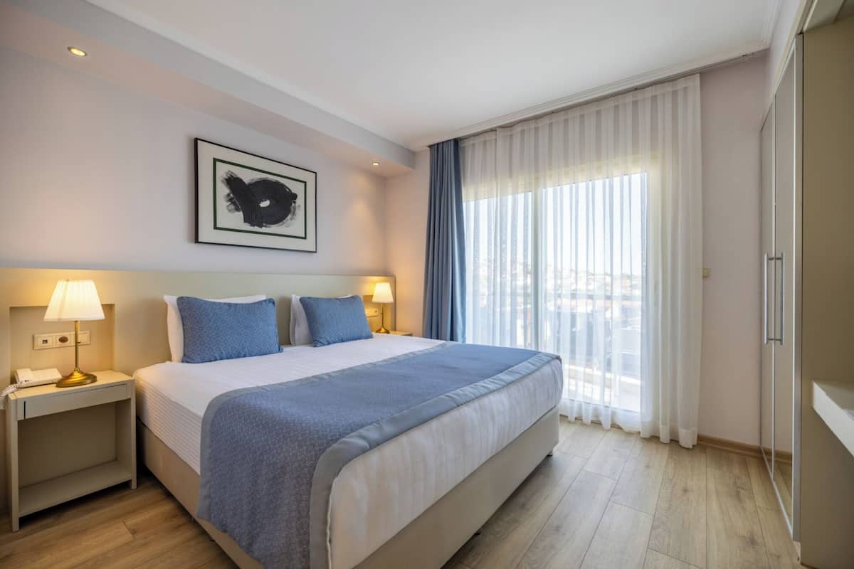A hotel room with a large bed, two bedside lamps, a framed artwork on the wall, and a view of the city through sheer curtains.