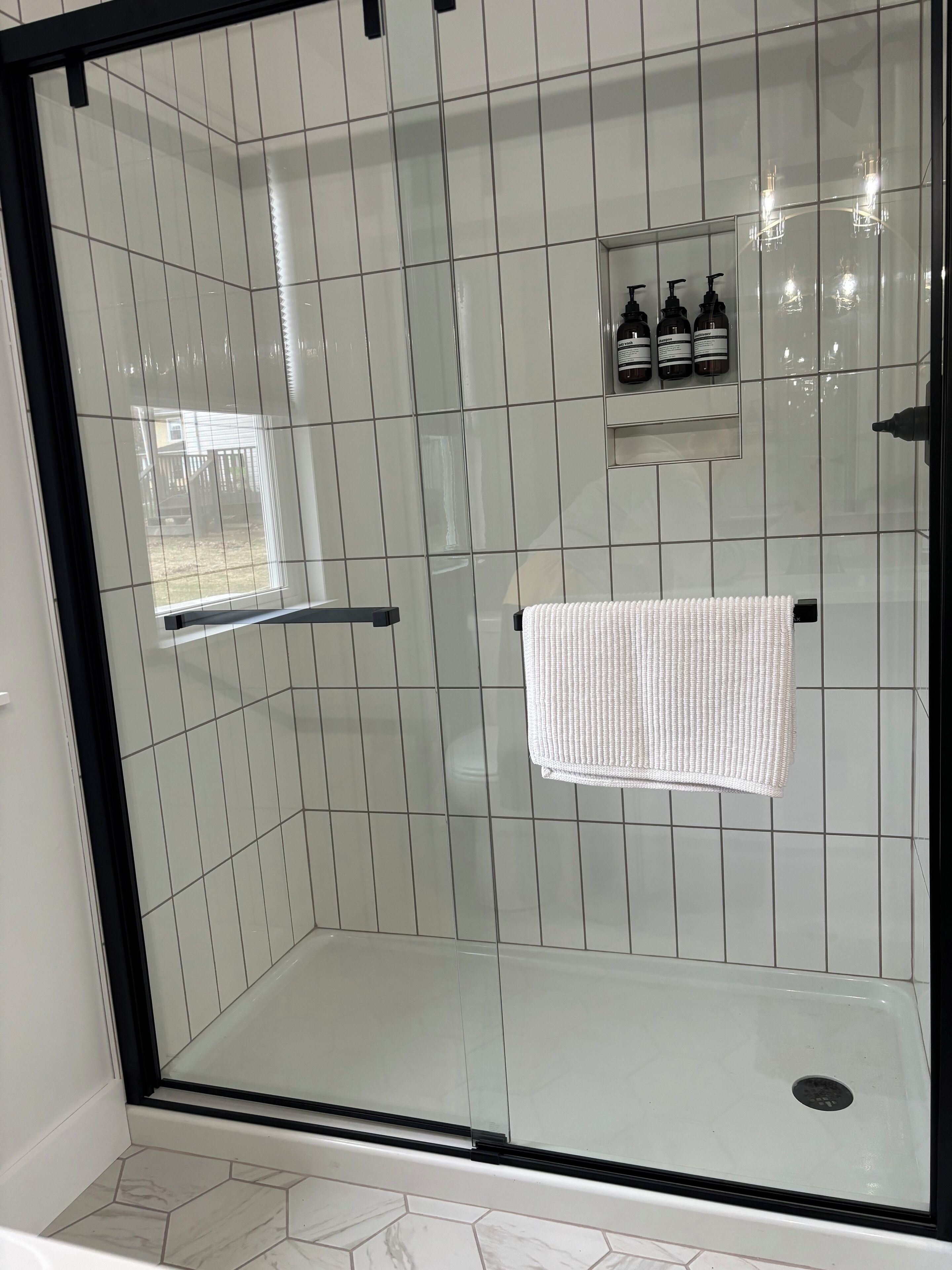 Une douche fermée par des parois de verre, avec une serviette blanche suspendue à un porte-serviettes, une petite étagère contenant des articles de toilette et un sol carrelé avec des carreaux hexagonaux.