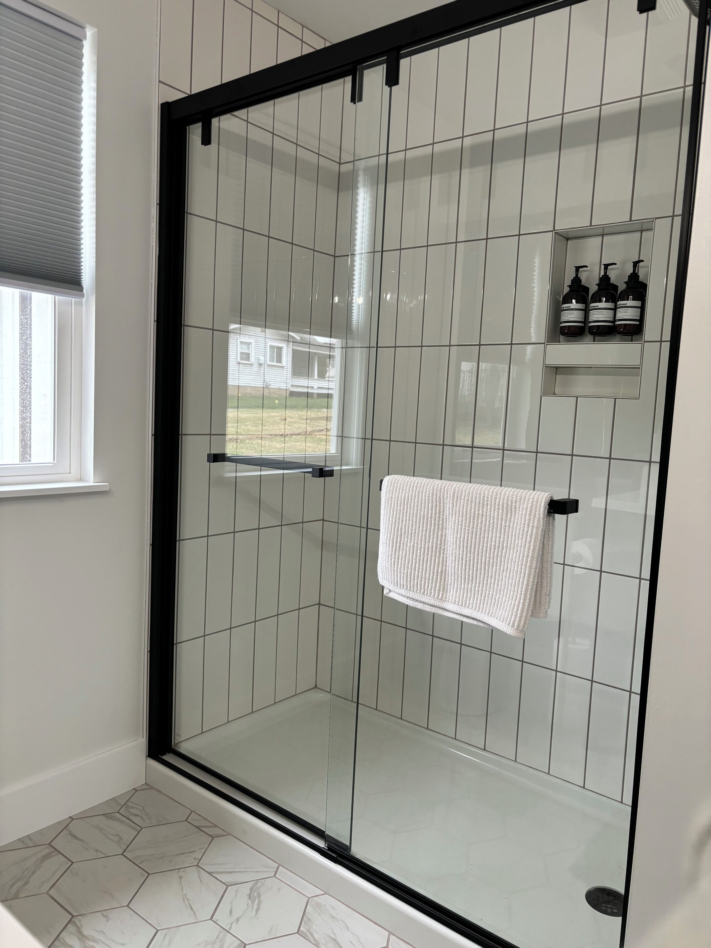 A glass-enclosed shower with a white towel hanging on a rack, hexagonal patterned floor tiles, and a small shelf with toiletries.