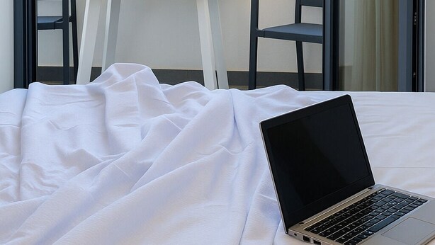 A bedroom with a laptop on a bed, a table with chairs, and a view of the sea and buildings. - Filia Skyros Suites (Skiros)