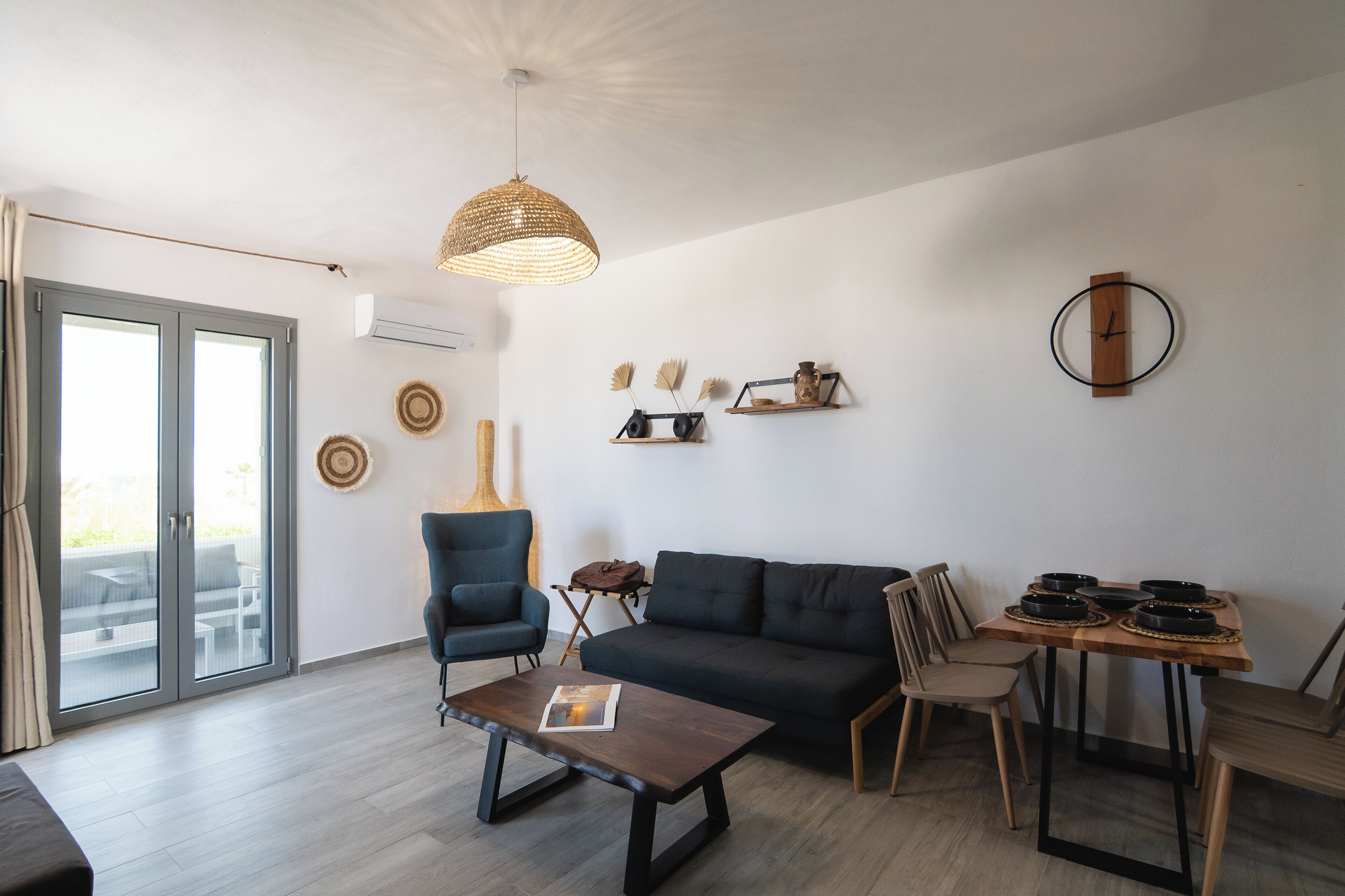 A modern living room with a sofa, chairs, a coffee table, and a dining table. There is a large window with curtains, a hanging light fixture, and wall decorations.