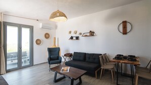 A modern living room with a sofa, chairs, a coffee table, and a dining table. There is a large window with curtains, a hanging light fixture, and wall decorations. - Filia Skyros Suites (Skiros)